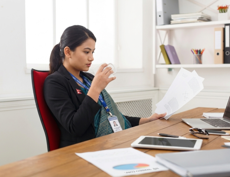 A professional Nepali woman in a black blazer with an ID badge reviews documents while drinking tea at her office desk, with a laptop, tablet, and charts nearby.